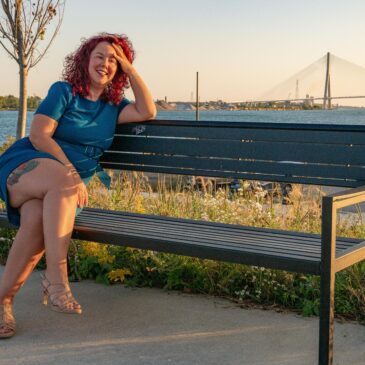 Bridget Espinosa sitting casually on a bench with the Gordie Howe International Bridge in the background with wind blown red hair.