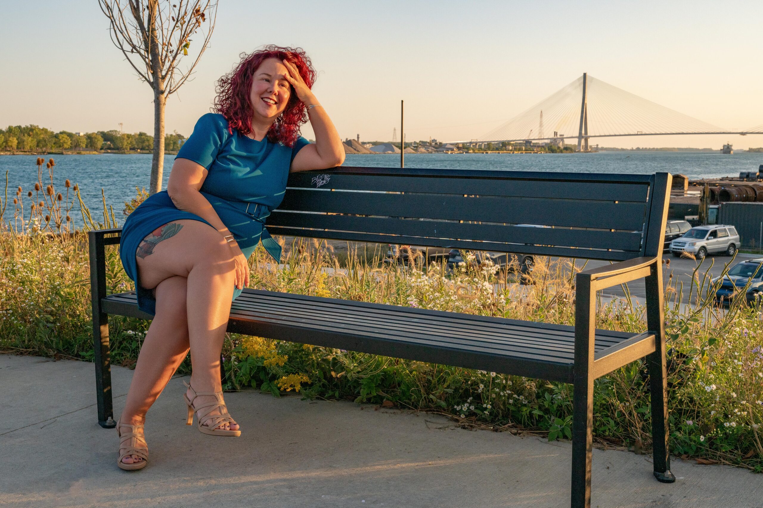 Bridget Espinosa sitting casually on a bench with the Gordie Howe International Bridge in the background with wind blown red hair.